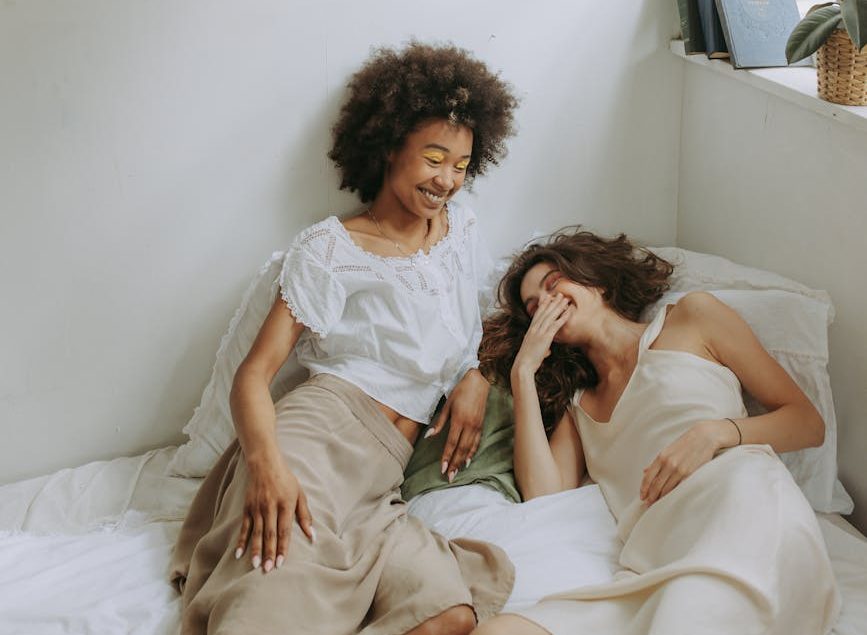 two women talking and laughing while sitting on the bed